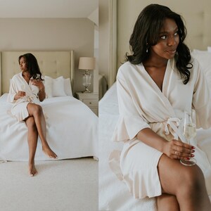 May include: A cream-colored robe with a tie waist and ruffled sleeves. The robe is worn by a person sitting on a bed with white linens. The room has neutral tones and is lit by natural light. A glass of champagne is held.