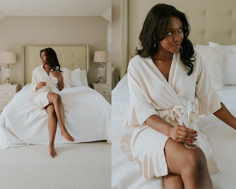 May include: A cream-colored robe with a tie waist and ruffled sleeves. The robe is worn by a person sitting on a bed with white linens. The room has neutral tones and is lit by natural light. A glass of champagne is held.