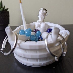 May include: White ceramic bath tub with a figurine of a woman inside. The tub is filled with blue and white beads, a pencil, and a few dice. There are three silver rings hanging on the side of the tub.