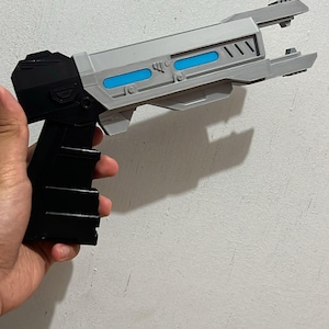May include: A sci-fi style toy blaster featuring a black handle and base with a light gray barrel. The barrel includes two horizontal blue rectangular accents and a pronged front tip. The design has a geometric, angular construction with visible panel lines.