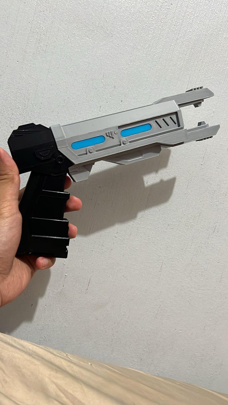 May include: A sci-fi style toy blaster featuring a black handle and base with a light gray barrel. The barrel includes two horizontal blue rectangular accents and a pronged front tip. The design has a geometric, angular construction with visible panel lines.