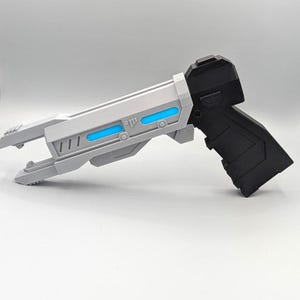 May include: A futuristic toy blaster featuring a matte black handle and a light gray barrel assembly. The barrel includes two horizontal recessed slots filled with bright cyan blue accents, along with geometric paneling and a split-prong front muzzle.