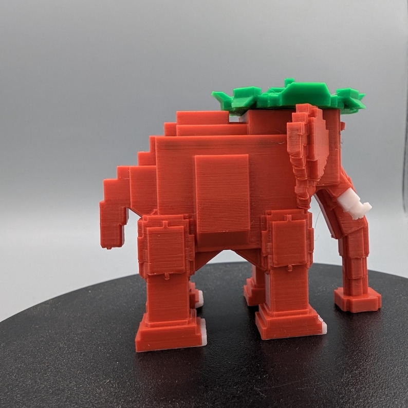 May include: A 3D-printed, pixelated elephant figurine in shades of red, with white tusks and feet, and a green leafy crown. The elephant is designed with a blocky, geometric aesthetic, giving it a retro video game appearance.