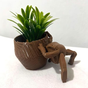 May include: A brown 3D-printed planter with a geometric textured pattern, featuring an attached articulated figure leaning against the side. The planter contains a small, vibrant green artificial plant with pointed leaves extending upwards.