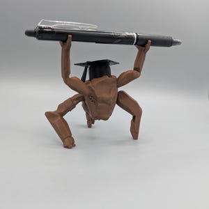 May include: A brown, articulated figurine wearing a graduation cap, holding a black pen overhead. The pen has a silver clip and a blue ink tip. The figurine has multiple arms and legs, and is posed as if lifting the pen.