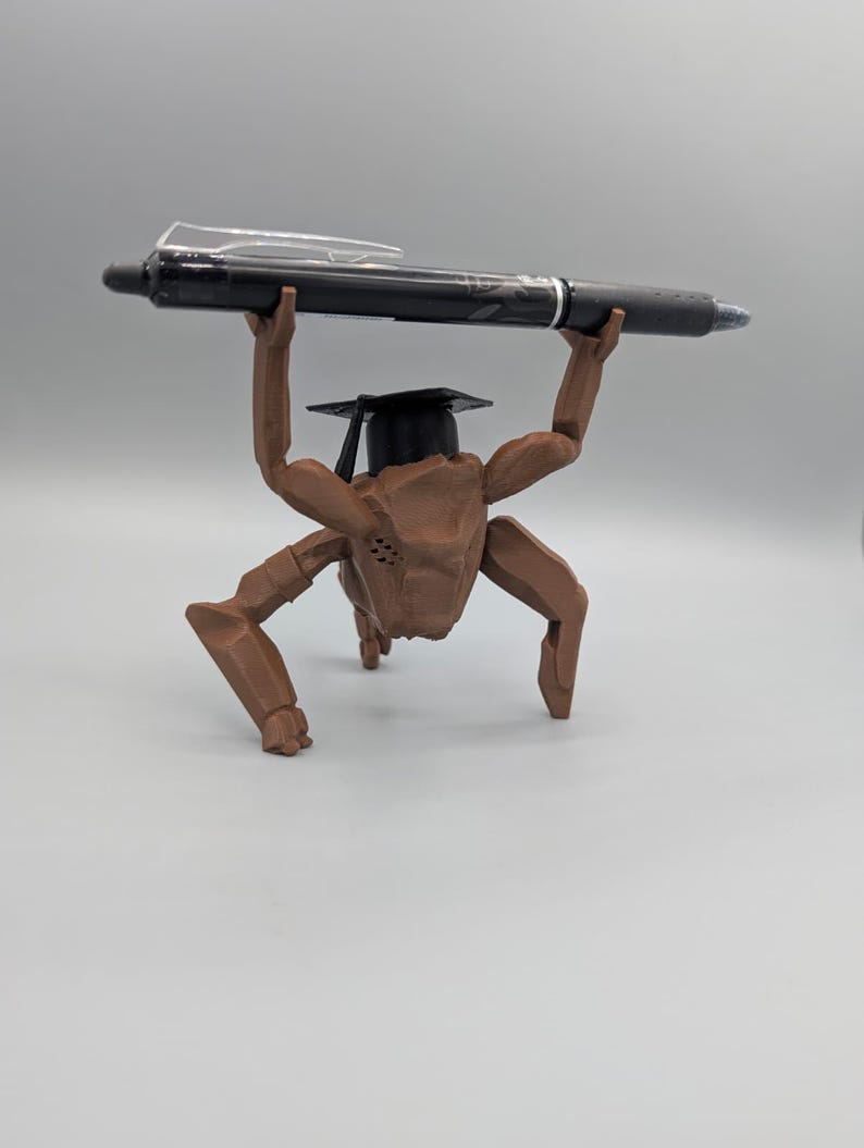 May include: A brown, articulated figurine wearing a graduation cap, holding a black pen overhead. The pen has a silver clip and a blue ink tip. The figurine has multiple arms and legs, and is posed as if lifting the pen.