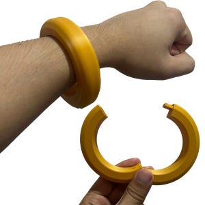 May include: A mustard yellow bangle bracelet, shown both worn and unclasped. The bracelet is a thick, rounded design, with a simple clasp mechanism. The image shows the bracelet on a person's wrist and in their hand.