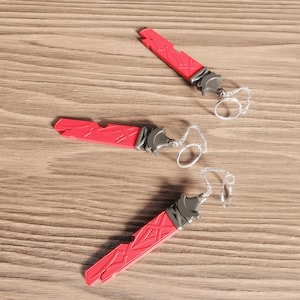 Puede incluir: Tres llaveros rojos con forma de espada. Cada llavero tiene un detalle metálico plateado y una pequeña cadena con un anillo. Los llaveros están colocados sobre una superficie de madera.