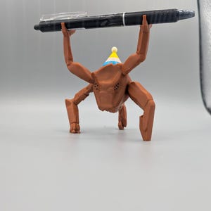 May include: A brown, insect-like figurine wearing a party hat, holding a black pen above its head. The pen has a clear clip and silver accents. The figurine has multiple legs and a detailed face, set against a plain background.