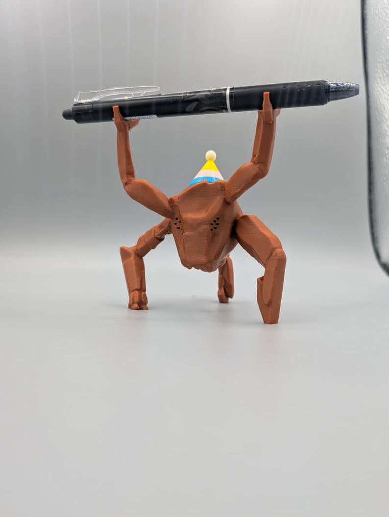 May include: A brown, insect-like figurine wearing a party hat, holding a black pen above its head. The pen has a clear clip and silver accents. The figurine has multiple legs and a detailed face, set against a plain background.