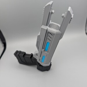 May include: A gray and black toy blaster with a futuristic design. The main body is light gray with two bright blue rectangular accents on the side. It features a black handle and a black base, with a wide, open-ended barrel structure at the top.