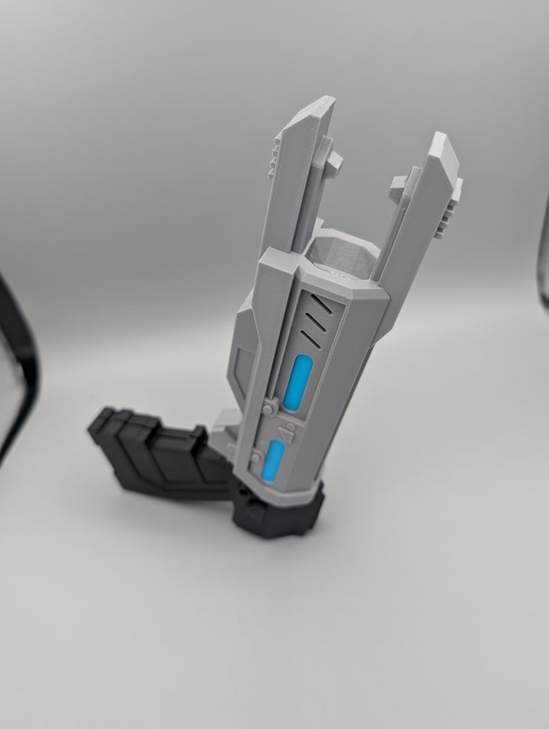 May include: A gray and black toy blaster with a futuristic design. The main body is light gray with two bright blue rectangular accents on the side. It features a black handle and a black base, with a wide, open-ended barrel structure at the top.