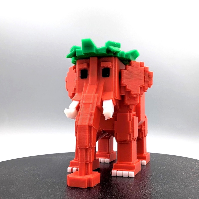May include: A red, pixelated elephant figurine with white tusks and a green leafy crown. The 3D-printed elephant has a blocky design, giving it a retro video game aesthetic. The figurine is standing on a dark surface.