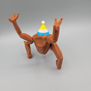 May include: A brown, articulated figurine with a party hat. The figurine has a geometric design and is posed with its arms raised. The party hat is yellow, blue, and white with a small white ball on top. The background is a solid gray.