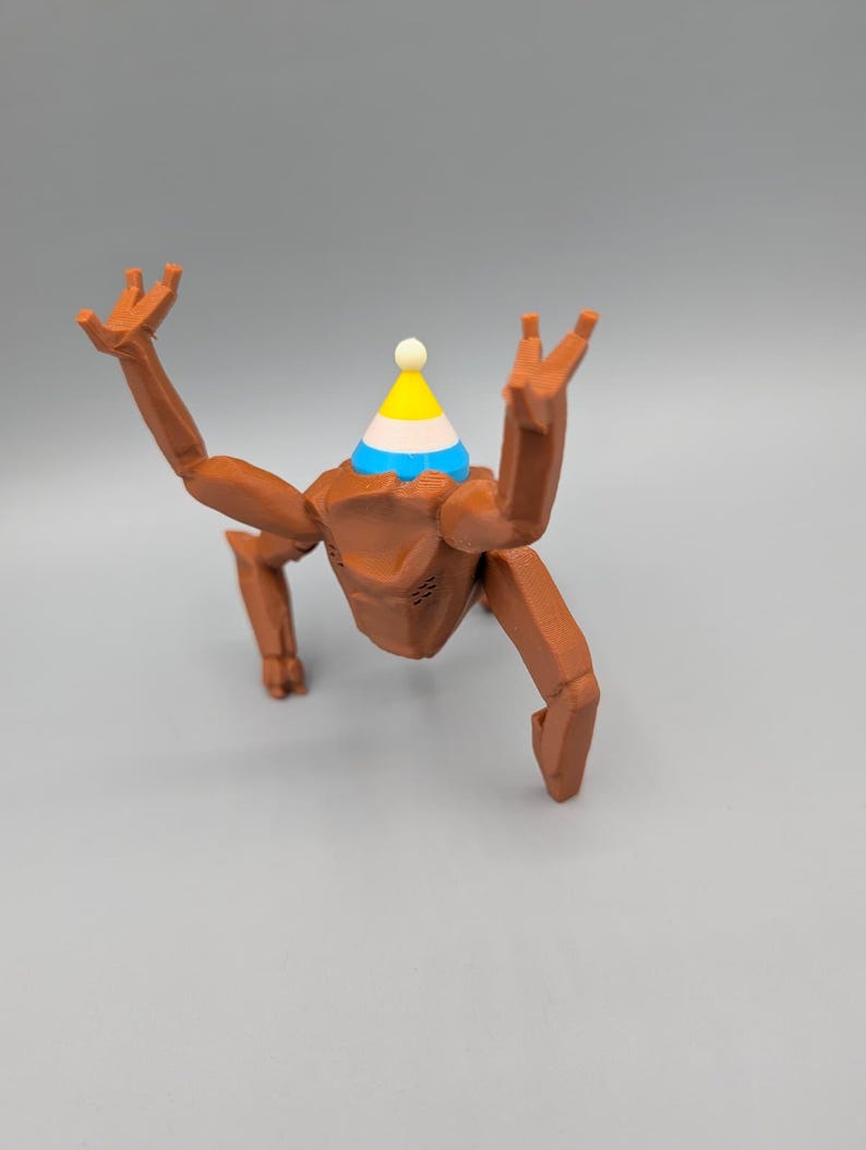 May include: A brown, articulated figurine with a party hat. The figurine has a geometric design and is posed with its arms raised. The party hat is yellow, blue, and white with a small white ball on top. The background is a solid gray.