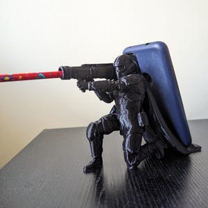May include: A black 3D printed phone stand featuring a kneeling action figure holding a weapon. The figure is holding a pencil in the weapon.
