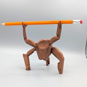 May include: A brown, multi-legged, creature-shaped desk organizer figurine with a textured surface. The figure is posed with its two upper limbs raised, balancing a long, orange Amazon Basics HB pencil horizontally across its hands.