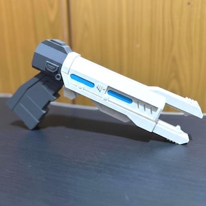 May include: A sci-fi style toy blaster featuring a dark gray handle and base, a white barrel assembly, and bright blue rectangular accents along the sides. The design includes geometric paneling, a trigger guard, and a split-prong muzzle end.