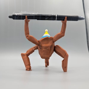 May include: A brown, insect-like figurine wearing a party hat, holding a black pen. The pen is held above the figurine's head. The figurine has multiple legs and arms. The party hat is blue, yellow, and white.