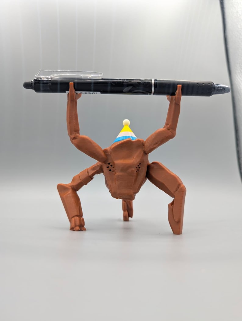 May include: A brown, insect-like figurine wearing a party hat, holding a black pen. The pen is held above the figurine's head. The figurine has multiple legs and arms. The party hat is blue, yellow, and white.