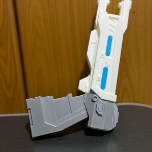 May include: A futuristic toy blaster featuring a white barrel with two bright blue rectangular accents and a gray handle section. The object has a rigid, angular construction with visible layered textures typical of a 3D printed item.