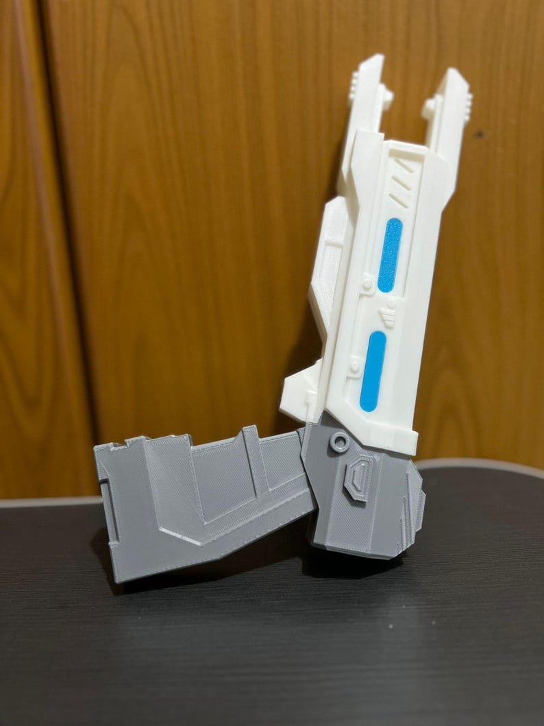 May include: A futuristic toy blaster featuring a white barrel with two bright blue rectangular accents and a gray handle section. The object has a rigid, angular construction with visible layered textures typical of a 3D printed item.