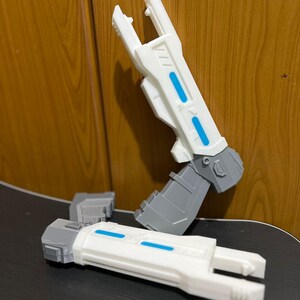 May include: Two futuristic toy blasters featuring a white barrel with two horizontal blue rectangular accents and a gray handle. One blaster is positioned vertically, while the other lies horizontally, both showcasing a geometric, angular design.