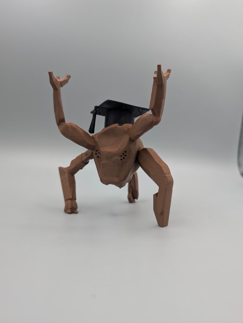 May include: A brown, stylized, four-legged creature with raised arms and a black graduation cap. The figure has a textured surface and is posed against a plain background, suggesting a whimsical or academic theme.
