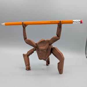 May include: A brown, stylized creature figurine holding an orange pencil with the text "amazon basics" printed on it. The pencil has a pink eraser. The figurine has multiple legs and arms, and a detailed head.