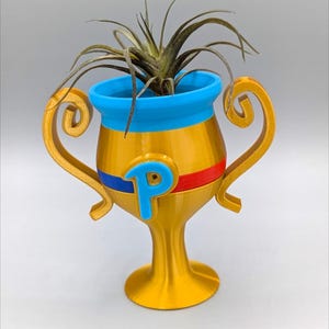 May include: A gold trophy-shaped planter with blue and red accents. The planter has a blue "P" on the front and is filled with a green air plant.
