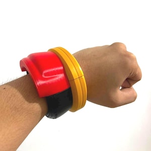 May include: A wide, multi-layered cuff bracelet featuring a bright red top section, a solid black middle band, and a thick yellow outer rim. The accessory has a smooth, rigid construction and wraps around the wrist with a bold, layered design.