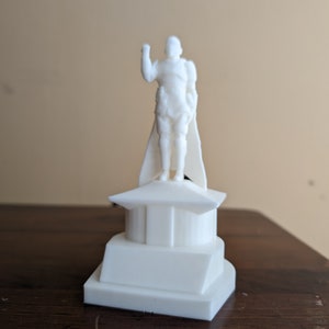 Könnte beinhalten: Weiße 3D-gedruckte Figur eines Menschen, der auf einer weißen Plattform mit quadratischem Sockel steht. Die Person trägt einen Umhang und hat den rechten Arm erhoben.