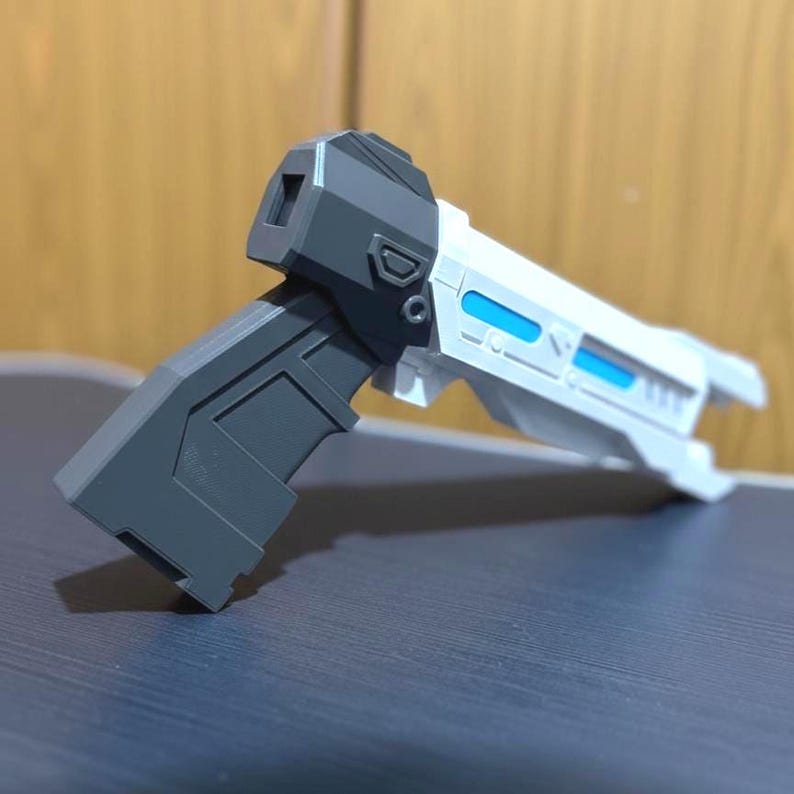 May include: A futuristic toy blaster featuring a dark gray handle and a white barrel assembly. The barrel includes two rectangular bright blue accents along the side, with a dark gray housing near the grip and a square opening at the rear.