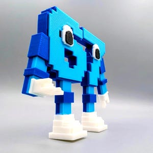 May include: A 3D-printed, pixelated robot figure in shades of blue and white. The robot has a blocky design with a head, arms, legs, and feet. The eyes are black and white. The figure is standing on a gray surface.