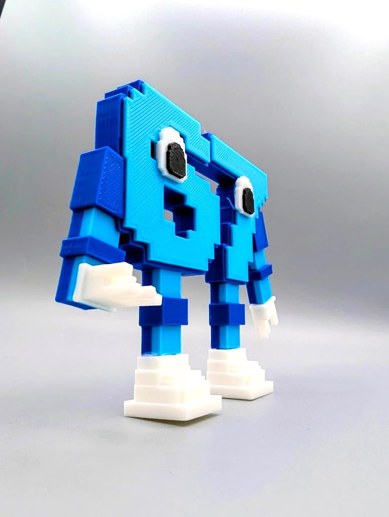 May include: A 3D-printed, pixelated robot figure in shades of blue and white. The robot has a blocky design with a head, arms, legs, and feet. The eyes are black and white. The figure is standing on a gray surface.