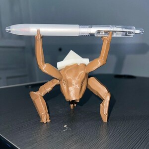 May include: A brown, 3D-printed creature figure holding a clear and white pen. The figure has multiple legs and a white headpiece. The pen is held above the figure's head. The figure is on a dark surface.