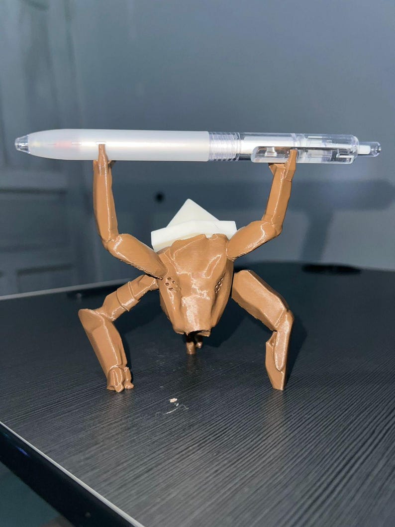 May include: A brown, 3D-printed creature figure holding a clear and white pen. The figure has multiple legs and a white headpiece. The pen is held above the figure's head. The figure is on a dark surface.