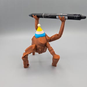 May include: A brown 3D-printed figurine with a party hat, holding a black pen. The pen has white text that reads "removed by friction". The figurine has a unique, abstract design.