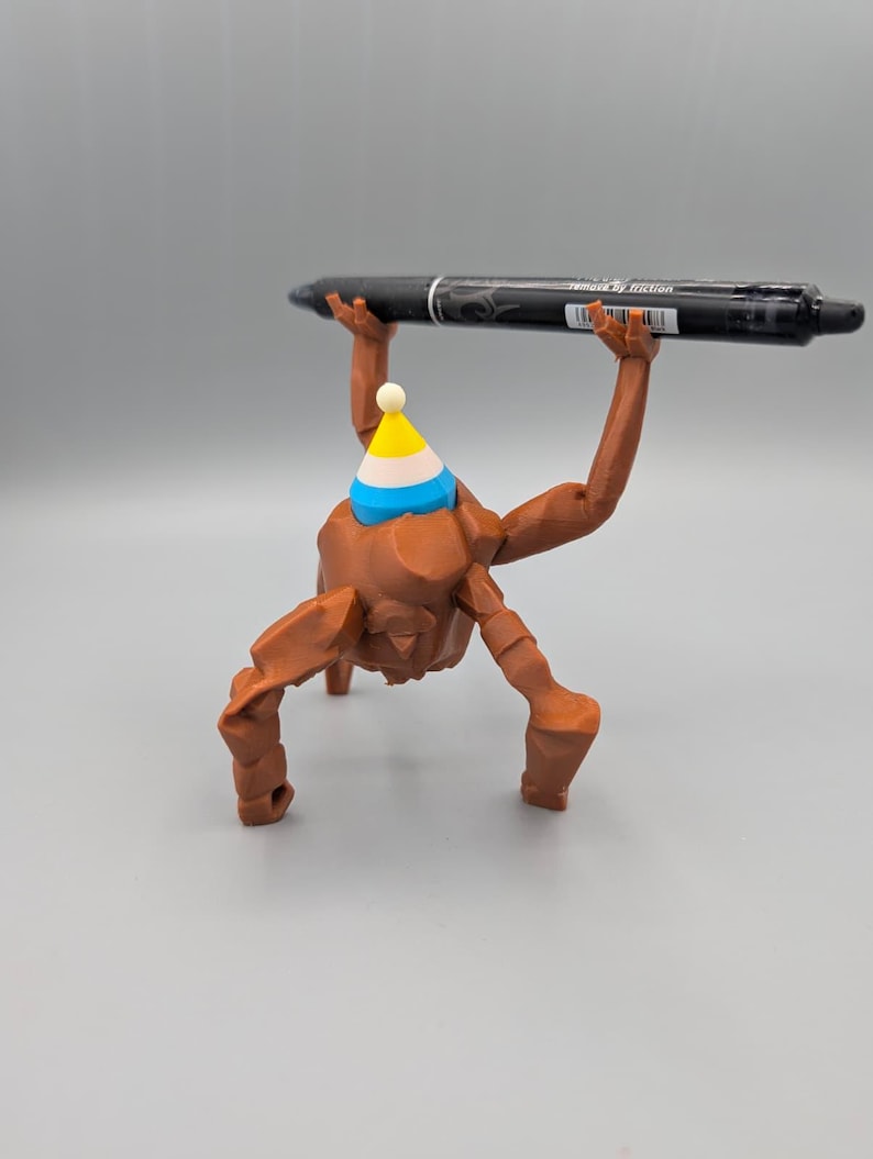 May include: A brown 3D-printed figurine with a party hat, holding a black pen. The pen has white text that reads "removed by friction". The figurine has a unique, abstract design.
