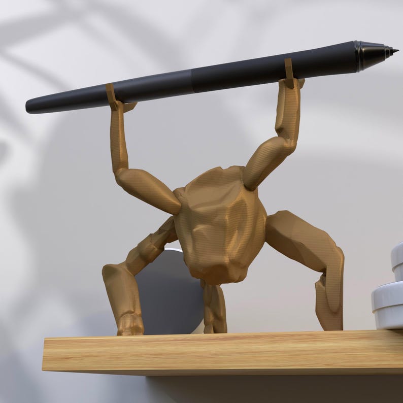 May include: A gold-colored pen holder in the shape of a stylized figure, holding a black pen. The pen holder is on a wooden shelf. The pen is black with a silver tip.