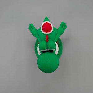 Puede incluir: Una figura verde de Yoshi con un ojo rojo y blanco y un cuerpo verde. La figura tiene los brazos extendidos y un hocico verde redondo. El fondo es gris liso.