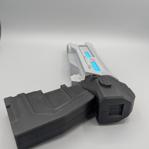 May include: A futuristic toy blaster featuring a matte black handle and a silver barrel assembly. The barrel section includes two rectangular cutouts with bright blue glowing inserts. The design has sharp, angular edges and a blocky, geometric structure.