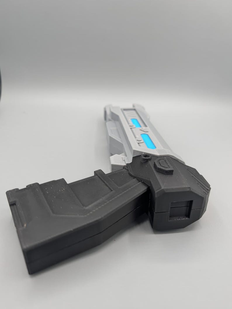 May include: A futuristic toy blaster featuring a matte black handle and a silver barrel assembly. The barrel section includes two rectangular cutouts with bright blue glowing inserts. The design has sharp, angular edges and a blocky, geometric structure.