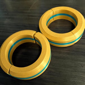May include: Two circular, two-piece rings featuring a matte gold exterior and a vibrant cyan blue inner band. Each ring is constructed from two interlocking halves, creating a distinct seam where the segments meet, forming a solid, uniform circular shape.