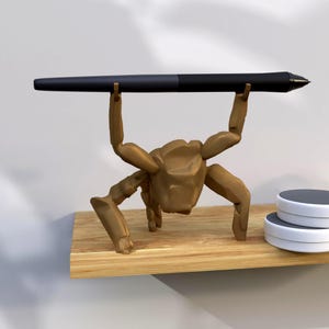 May include: A brown, stylized figure holds a black stylus pen aloft. The figure is positioned on a wooden shelf, with two white and gray coasters visible to the right. The background is a plain white wall.
