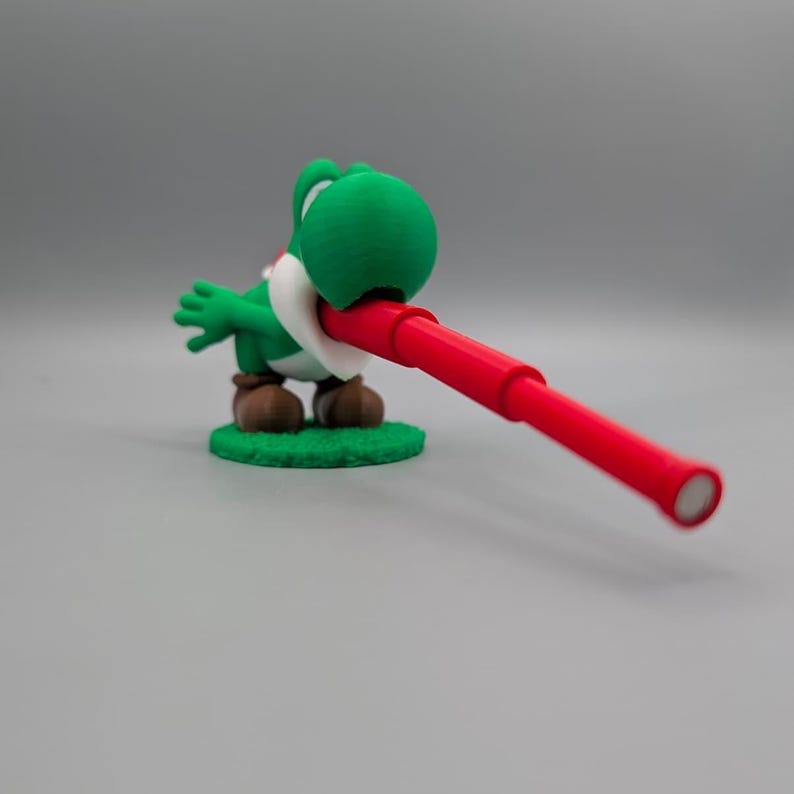Puede incluir: Una figura verde de Yoshi sosteniendo un telescopio rojo. La figura est&aacute; sobre una peque&ntilde;a base verde. Yoshi tiene un pecho blanco y zapatos marrones. El telescopio rojo est&aacute; inclinado hacia la derecha.