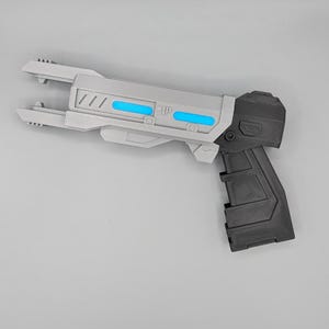 May include: A sci-fi style toy blaster featuring a light gray barrel with two horizontal bright blue accents and a dark gray handle. The barrel has a pronged front end and geometric detailing along the side, while the grip has a textured surface.