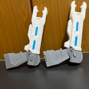 May include: Two white and gray plastic toy blasters featuring a futuristic design with elongated barrels, angular grips, and two bright blue rectangular accents on the side of each barrel. The blasters have a mechanical aesthetic with visible panel lines.
