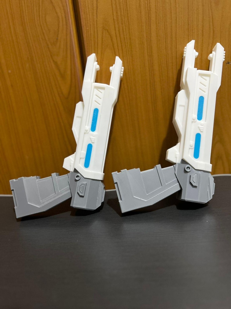 May include: Two white and gray plastic toy blasters featuring a futuristic design with elongated barrels, angular grips, and two bright blue rectangular accents on the side of each barrel. The blasters have a mechanical aesthetic with visible panel lines.