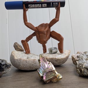 May include: A brown 3D-printed insect-like sculpture holding a blue and black marker labeled "PC-5M". The sculpture is positioned on a stone, with colorful crystals and other stones nearby. The background is a light-colored wall.